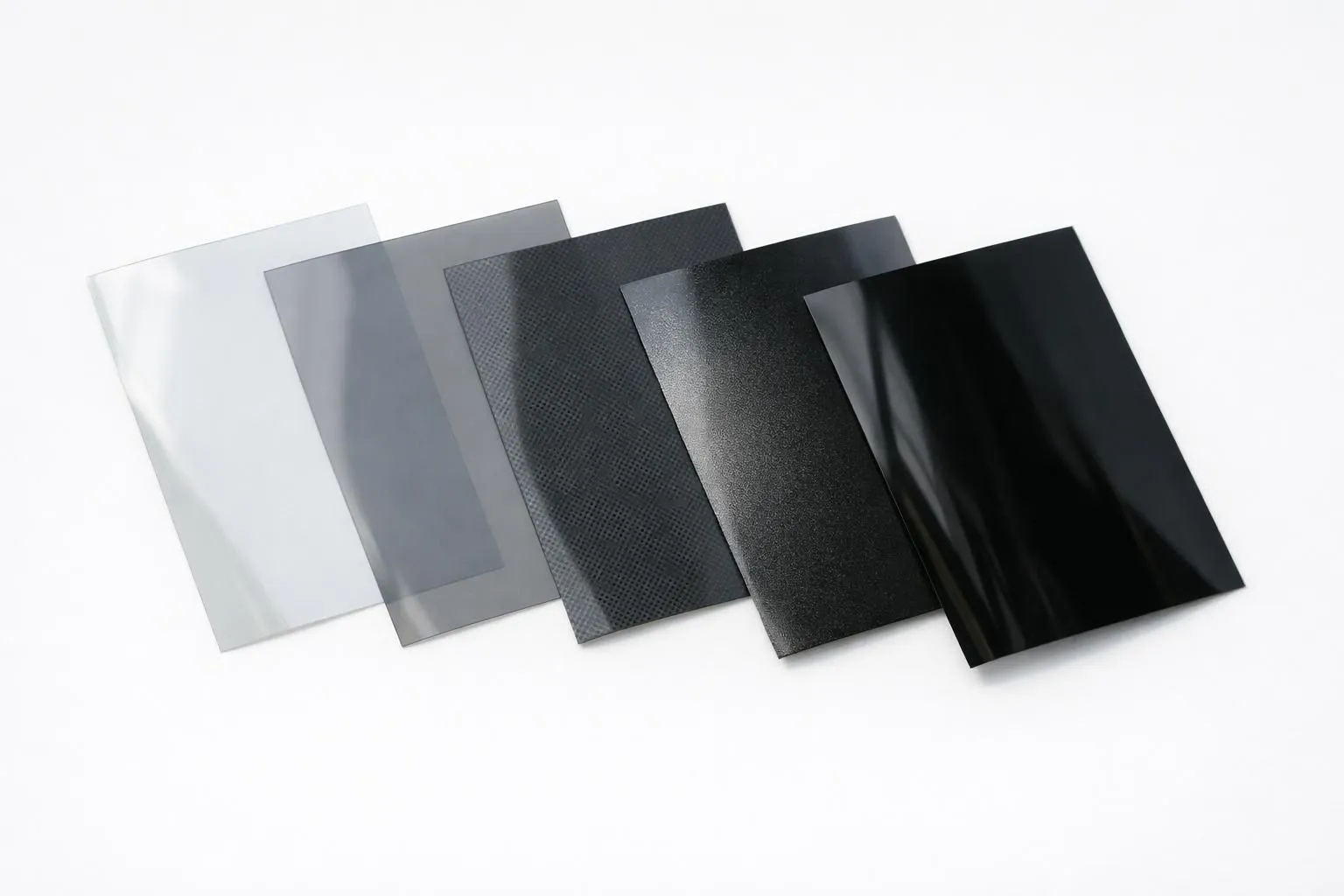 Cinq échantillons de films de teintage de fenêtre automobile disposés sur une surface blanche, montrant des dégradés de teintes claires à foncées avec des différences de texture visibles entre les finitions céramique, carbone et métallique, photographie professionnelle de produit avec éclairage naturel